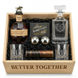 Blanton's Bourbon Date Night gift crate top-down view featuring "BETTER TOGETHER" engraved on front of open wooden crate, Blanton's Original Single Barrel Bourbon 750ml with brass horse stopper, Better Together square crystal decanter with pointed shield crest showing two intertwined hearts, "BETTER TOGETHER" italic text, and elaborate baroque acanthus leaf scrollwork flourishes, two XXL 55mm polished chrome stainless steel whiskey balls, two old-fashioned crystal glasses, and Turin chocolates.