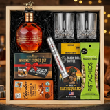 Blanton's Gold Edition Single Barrel Bourbon 700ml gift crate with gold horse stopper, crystal-cut old fashioned glasses, Royal Reserve whiskey stones set, Montecristo Platinum cigar, Black Rifle Coffee, Chocolove chocolate, and Wonderful Pistachios on rustic aged oak surface with warm amber bourbon bar lighting and moody bokeh