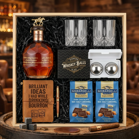 Blanton's Gold Edition Single Barrel Bourbon gift crate with gold horse stopper, crystal-cut old fashioned glasses, faux-leather bourbon journal and pen set, premium XXL stainless steel whiskey balls, and two Ghirardelli Dark Chocolate Sea Salt Caramel bags on rustic aged oak surface with warm amber bourbon bar lighting and moody bokeh