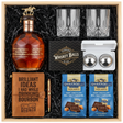 Blanton's Gold Edition Single Barrel Bourbon gift crate with gold horse stopper, crystal-cut old fashioned glasses, faux-leather bourbon journal and pen set, premium XXL stainless steel whiskey balls with freezer tray, and two Ghirardelli Dark Chocolate Sea Salt Caramel bags in black crinkle paper on white background