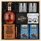 Blanton's Gold Edition Single Barrel Bourbon gift crate with gold horse stopper, crystal-cut old fashioned glasses, faux-leather bourbon journal and pen set, premium XXL stainless steel whiskey balls with freezer tray, and two Ghirardelli Dark Chocolate Sea Salt Caramel bags in black crinkle paper on white background