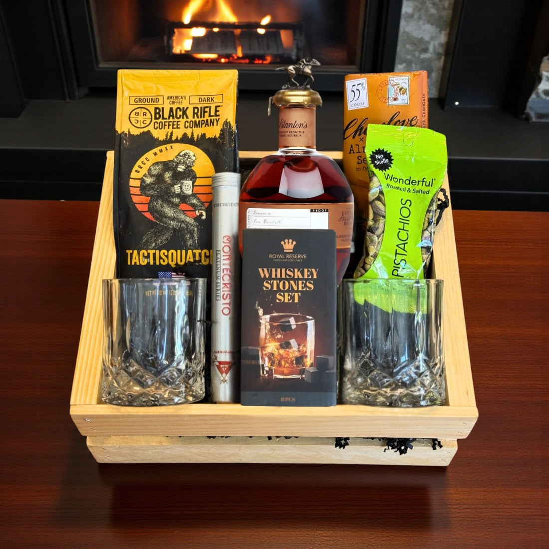 Engravable Blanton’s Straight From the Barrel Bourbon Gift Crate with coffee, chocolate, pistachios, cigar, whiskey stones, and glasses on wooden table near fireplace – Top Bourbon lifestyle image