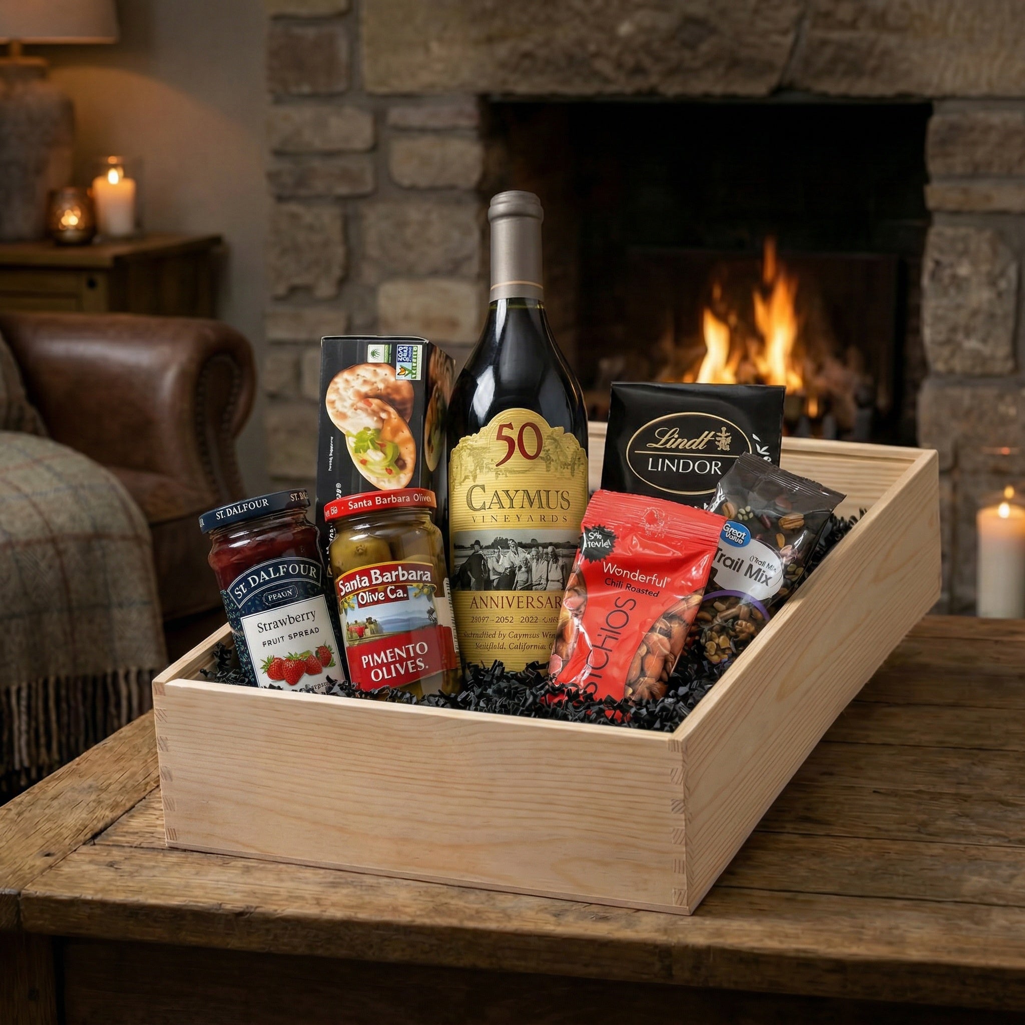 Caymus Vineyards Cabernet Sauvignon anniversary wine gift crate with Lindt Lindor chocolates, pistachios, olives, strawberry spread, crackers and trail mix displayed in wooden gift crate by fireplace