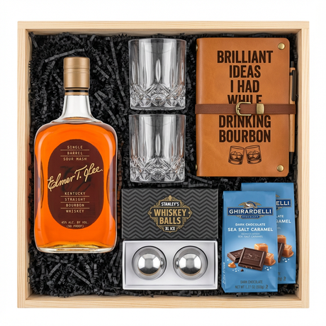 Bird's eye view of Elmer T. Lee Single Barrel Sour Mash Kentucky Straight Bourbon Whiskey gift crate with gold cap and dark brown oval label, crystal-cut old fashioned glasses, tan leather bourbon journal and pen set, Stanley's XXL stainless steel whiskey balls with rectangular freezer tray, and two Ghirardelli Dark Chocolate Sea Salt Caramel bags all nestled in black zigzag crinkle paper inside wooden crate on white background