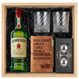Jameson Irish Whiskey gift crate with green bottle and red cap, crystal-cut old fashioned glasses with diagonal twisted swirl pattern, tan leather bourbon journal and pen set, and Stanley's XXL stainless steel whiskey balls all nestled in black zigzag crinkle paper inside wooden crate on white background