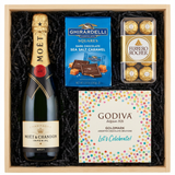 Moët & Chandon Impérial Brut Champagne gift crate with gold foil neck and red wax seal, Ghirardelli Dark Chocolate Sea Salt Caramel squares, Godiva Let's Celebrate chocolate box, and Ferrero Rocher chocolates in black crinkle paper on white background