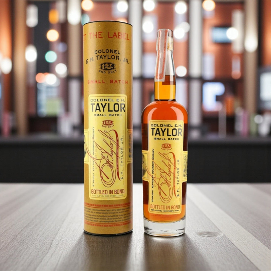 Engravable Colonel E.H. Taylor Small Batch Bourbon bottle and tube on wooden table with warm bar lighting – Top Bourbon lifestyle image