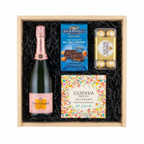 Veuve Clicquot Rosé Brut Champagne gift crate with Ghirardelli Dark Chocolate Sea Salt Caramel squares, Godiva Let's Celebrate chocolate box, and Ferrero Rocher chocolates in black crinkle paper on white background
