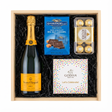 Veuve Clicquot Yellow Label Brut Champagne gift crate with Ghirardelli Dark Chocolate Sea Salt Caramel squares, Godiva Let's Celebrate chocolate box, and Ferrero Rocher chocolates in black crinkle paper on white background