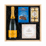 Veuve Clicquot Yellow Label Brut Champagne gift crate with Ghirardelli Dark Chocolate Sea Salt Caramel squares, Godiva Let's Celebrate chocolate box, and Ferrero Rocher chocolates in black crinkle paper on white background