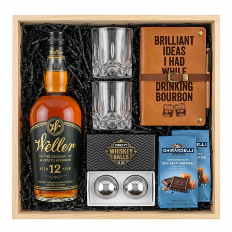 Bird's eye view of W.L. Weller 12 Year Wheated Bourbon gift crate with black label and gold text, crystal-cut old fashioned glasses, tan leather bourbon journal and pen set, Stanley's XXL stainless steel whiskey balls with rectangular freezer tray, and two Ghirardelli Dark Chocolate Sea Salt Caramel bags all nestled in black zigzag crinkle paper inside wooden crate on white background